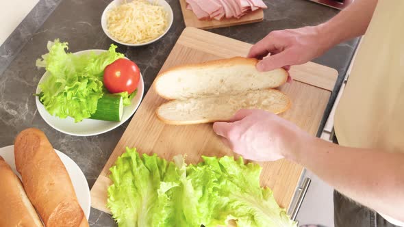 Hands spread lettuce leaves on a sandwich bun on a wooden cutting board.