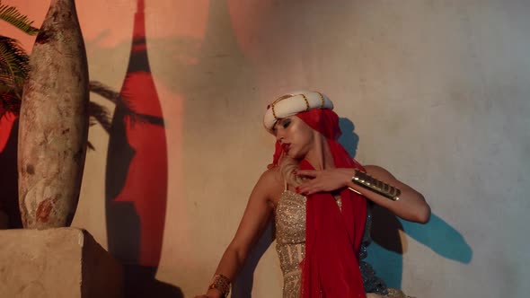 a Woman in a Turban with a Red Scarf and a Shiny Dress Sits on a Stone Staircase and Moves Hands alt