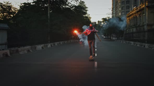 Young Fellow in Bandana on Face and Flag of USA Tied on His Chest is Riding Skateboard Along Street alt