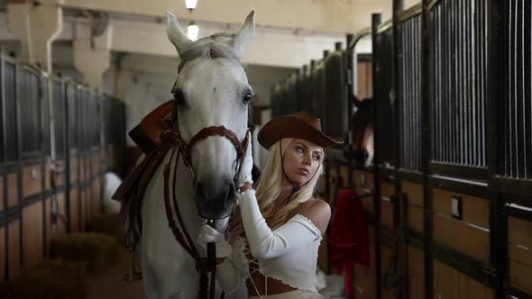 Portrait of a Blonde in a Cowboy Hat and with Big Breasts Next to a White Horse in the Stable alt