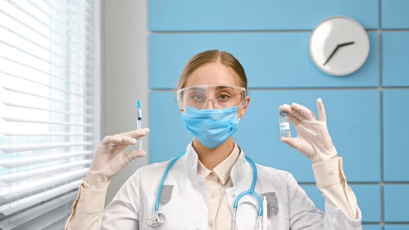 Young woman blonde in white coat and mask with stethoscope holds syringe alt