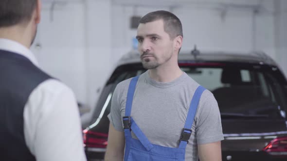 Portrait of Young Caucasian Auto Mechanic Talking with Customer in Repair Shop. Handsome Man  alt