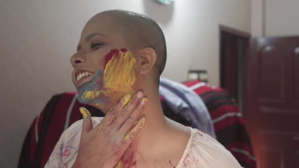 Happy and hopeful cancer patient bald girl applies colored powder on cheeks at home on Holi, Indian alt