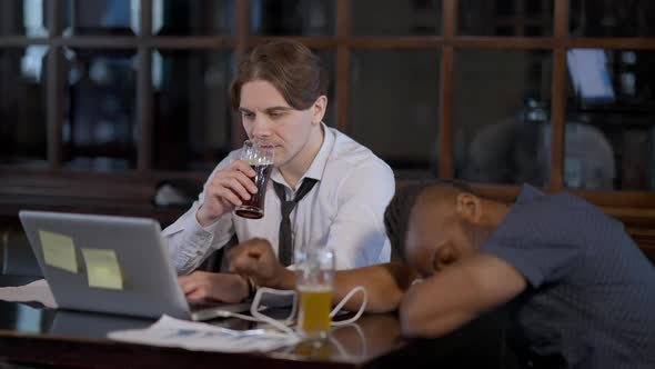 Confident Caucasian Man Working Online on Laptop in Restaurant Drinking Beer with Passed Out African alt