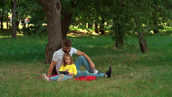 Father and Little Child Sit in Summer Park with Tablet