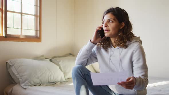 Mixed race woman working at home, sitting on bed talking on smartphone holding paperwork in cottage alt