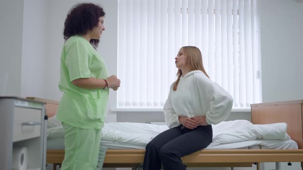 Young Woman Complaining Pain Sitting on Bed in Ward Talking to Nurse Standing in Hospital alt