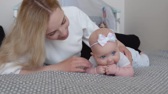 Baby Girl in Gray Clothes with a Cute Bow on His Head Lies Tummy Down on the Bed Near His Mother on alt