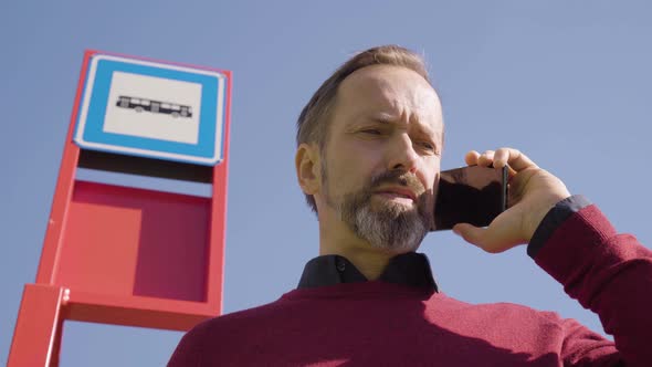 A Middleaged Handsome Caucasian Man Talks on a Smartphone at a Bus Stop alt