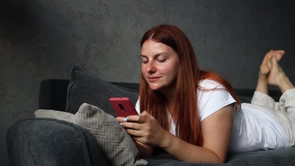 Redhaired Smiling Girl Lying on the Couch and Uses a Smartphone alt