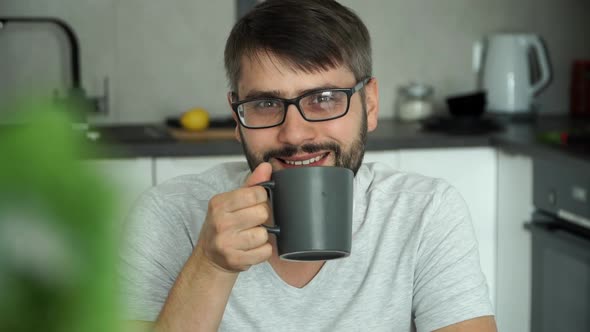 Portrait of Bearded Man Drinks Cup of Coffee Looks at Camera and Smiles at Home in Kitchen