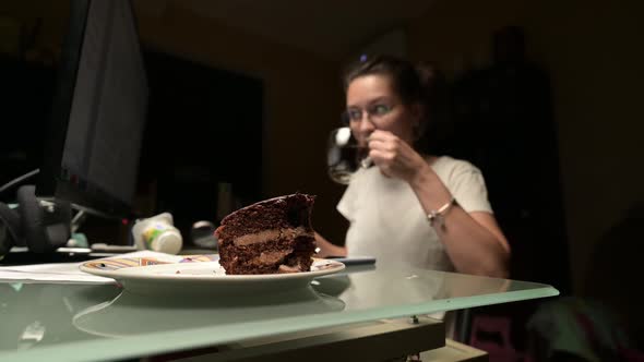 Young woman works at a computer at night with a cup of black coffee and a sweets alt