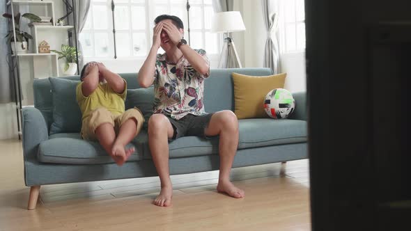 Frustrated Disappointed Asian Father And His Son Are Watching Tv While Sitting On A Couch At Home alt