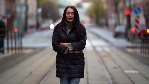 a Brunette with Long Hair in a Knitted Sweater and a Dark Coat Walks Along the Tram Tracks on a alt