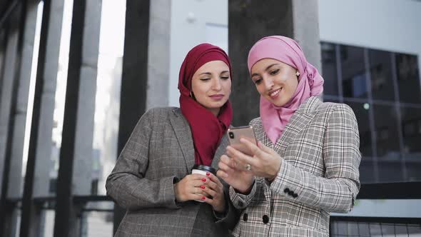 Two Young Muslim Women Wearing Hijab Headscarf Look at Phone, Walking Together Laughing Smile To