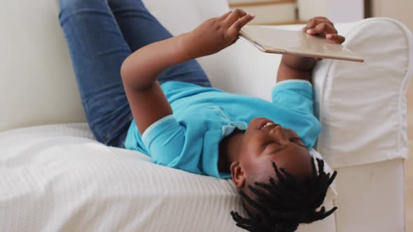 African american boy smiling while using digital tablet while lying on the couch at home alt