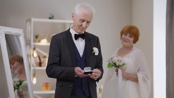 Proud Senior Caucasian Groom Posing with Wedding Rings As Happy Bride Standing at Background with alt