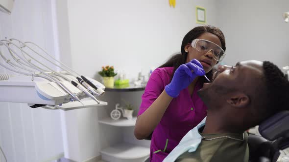 Beautiful Dark Skinned Woman Dentist in Pink Medical Siut and Blue Gloves Examines and Consults the alt