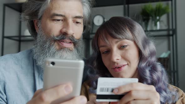 Joyful Husband and Wife Shopping Online Paying with Credit Card and Smartphone Laughing Indoors at alt