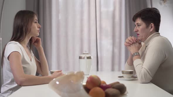 Close-up Side View of Caucasian Mother and Daughter Sitting at the Table with Tea Cups, Talking and alt
