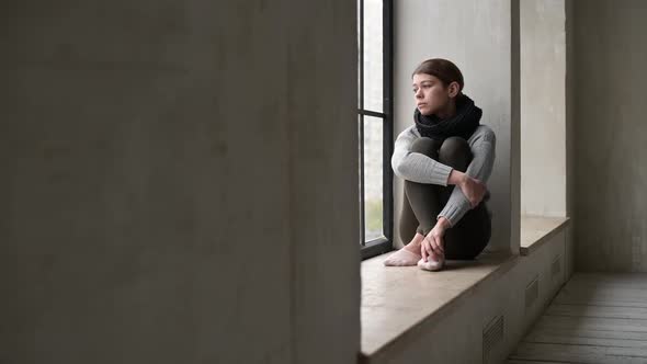 A sickly-looking young woman is sad on the windowsill of a city apartment alt