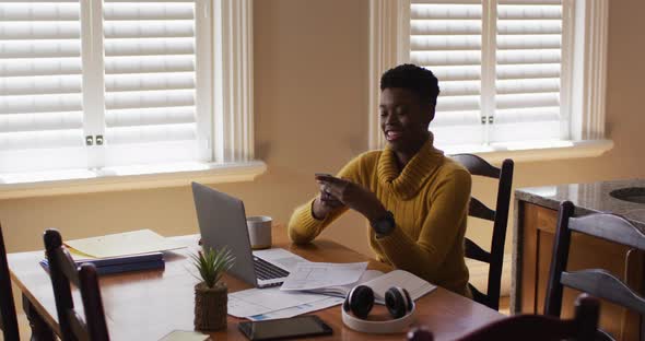 African american woman talking on smartphone and using laptop while working from home alt