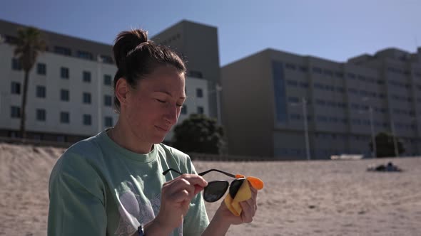 A Girl Sits on the Beach on the Sand Wiping Her Glasses and Putting Them on alt