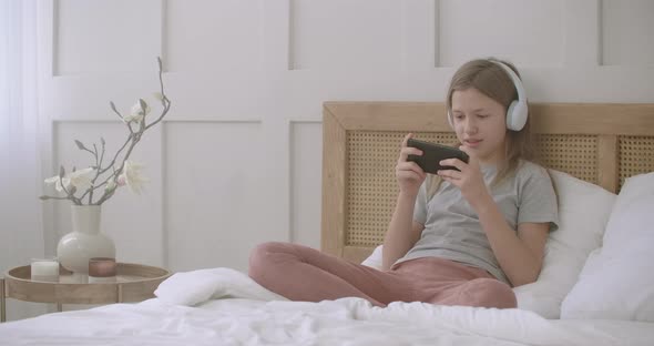Child Girl Is Playing Game in Smartphone After School Lessons, Sitting on Bed at Home Alone alt