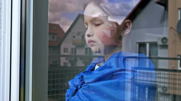 Little Girl with the Flag of Ukraine on Her Shoulders Looks Out the Window with a Serious Expression
