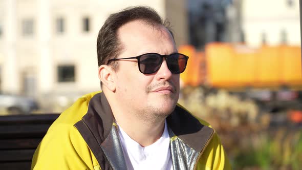 Portrait of Mature Handsome Guy in Sunglasses Sitting on Bench in City Park Looking Aside alt