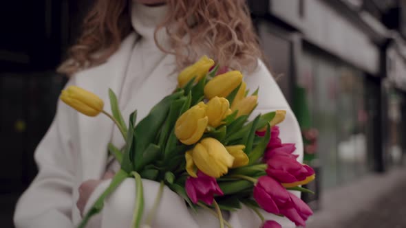 Nice Girl in White Coat with Bouquet of Tulips is Walking Down Street