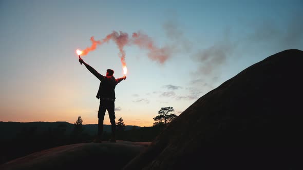 A Man is Standing on the Edge of a Cliff and Waving Two Flares in the Night alt