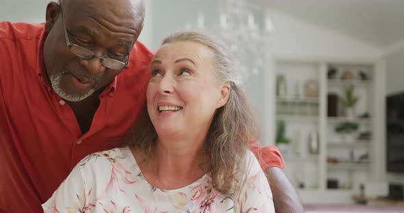 Happy senior diverse couple wearing shirts and embracing in living room alt