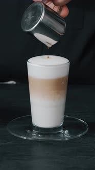 Barista pouring milk in hot coffee and making cappuccino foam in cafe close up, vertical alt