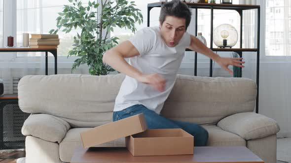 Caucasian Millennial Adult Man Buyer Consumer Receives Parcel Sitting on Couch Opening Cardboard Box alt