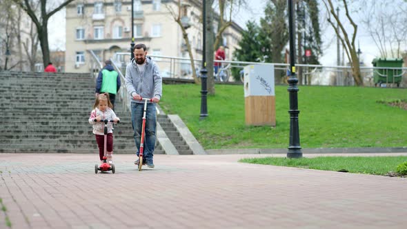 Happy Father and Little Daughter Riding Scooters in Park