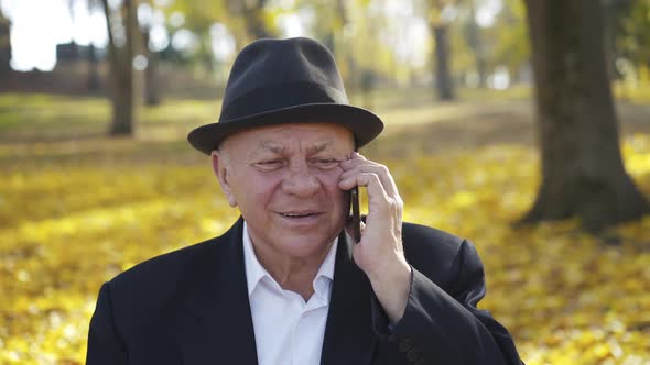 Senior Man in Hat Takes the Call and Has Fun Talks on the Mobile Phone on Bench alt