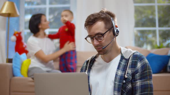 Businessman in Headset Having Video Call on Laptop While Wife Playing with Son on Background alt