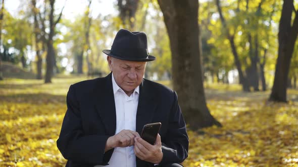 Thoughtful Senior Man in Suit and Hat Sits on Bench and Uses Smartphone in Park alt