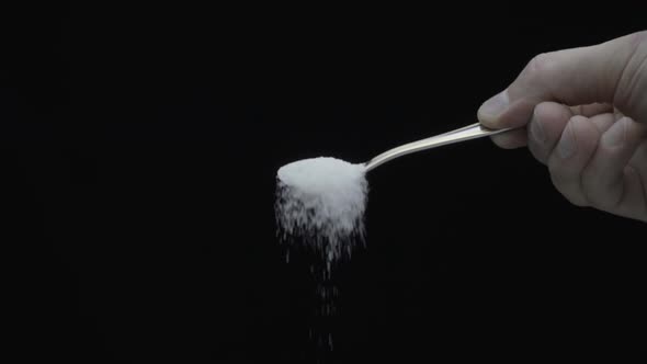 White Sugar From Teaspoon Fall Down On A Black Background. Slow Mo