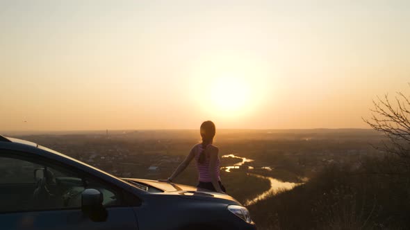 Young woman standing near her car enjoying warm sunset view. Girl traveler leaning alt