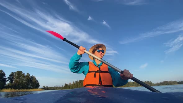 Young Man in Hat and Life Vest Rows Paddle Sitting in Boat alt