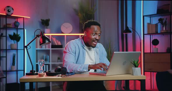 African American Sitting at the Desk in front of Laptop in Night Lighting Room alt
