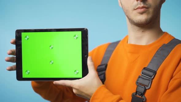 Young Man in Working Clothes Holding a Tablet with Green Screen and Smiling alt