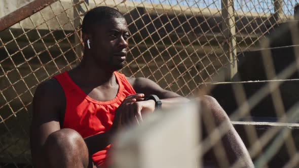 African american man sitting, checking his smartwatch, taking break in exercise outdoors alt