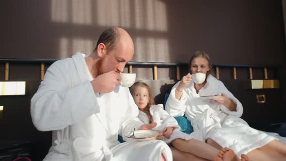 A Family in White Bathrobes Sitting in the Bed and Drinking Tea alt