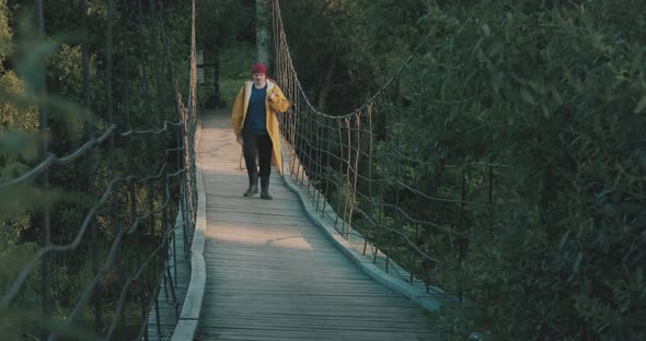 Man Hiking Tourist in Yellow Raincoat Walking Along Wooden Suspension Bridge alt