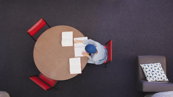 Asian female student wearing a blue hijab sitting at a round table and reading a book alt