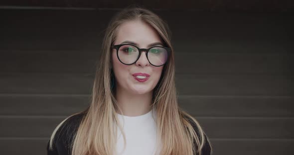 Stylish Teenager with Makeup in Glasses Inflates Chewing Gum Into the Camera alt