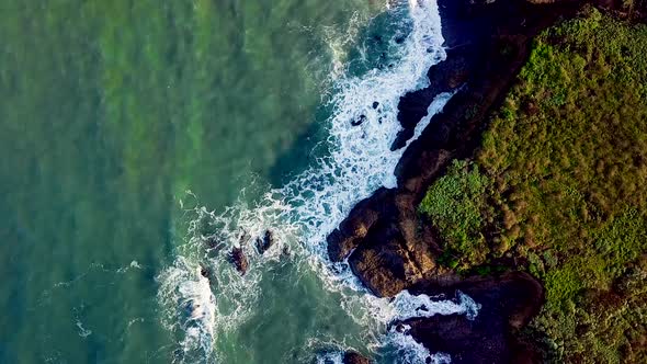 Aerial shot turning above sunlit waves crashing into rocky California cliffs alt